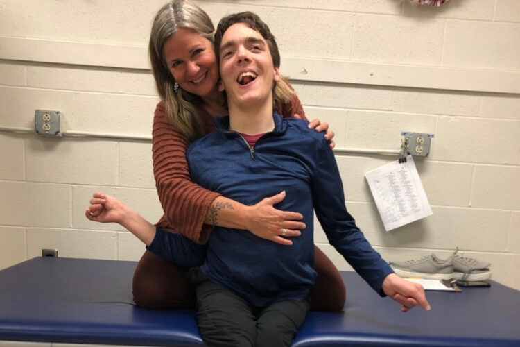 A blonde woman in her late forties embraces a young man with dark curly hair and long slightly misaligned limbs as they sit under tinsel on a bench and smile at the camera together.