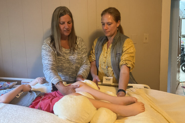 A blonde pale-skinned woman in her mid-forties shows another woman how to position the legs of a child who lies among a pile of therapeutic pillows.