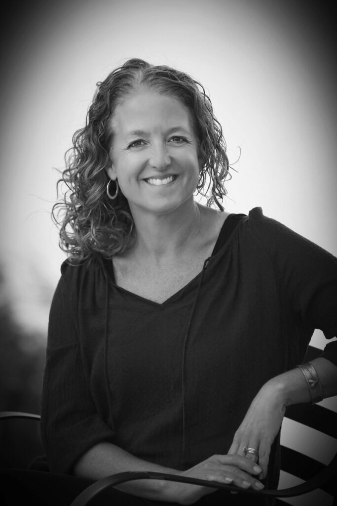 Black-and-white photo of a woman in her mid-forties with curly hair worn loose to her shoulders, wearing a black v-necked blouse and smiling casually at the camera