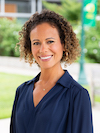 An almond-skinned woman in her early forties with walnut-brown hair in spiral curls, worn loose to her ears, wearing an open-collared navy blouse, and smiling directly at the camera