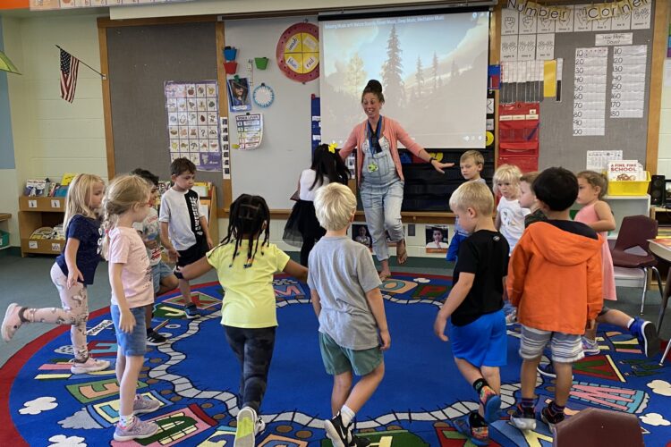 A pale-skinned teacher with medium-brown hair up in a messy bun and overalls stands on one foot in a brightly colored classroom. Around her a circle of students imitates her move.