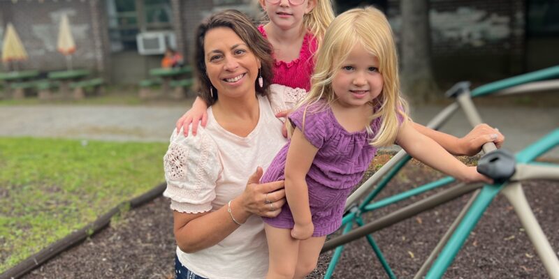 A pale-skinned woman holds two small blonde children who are climbing on a play dome in a playground.