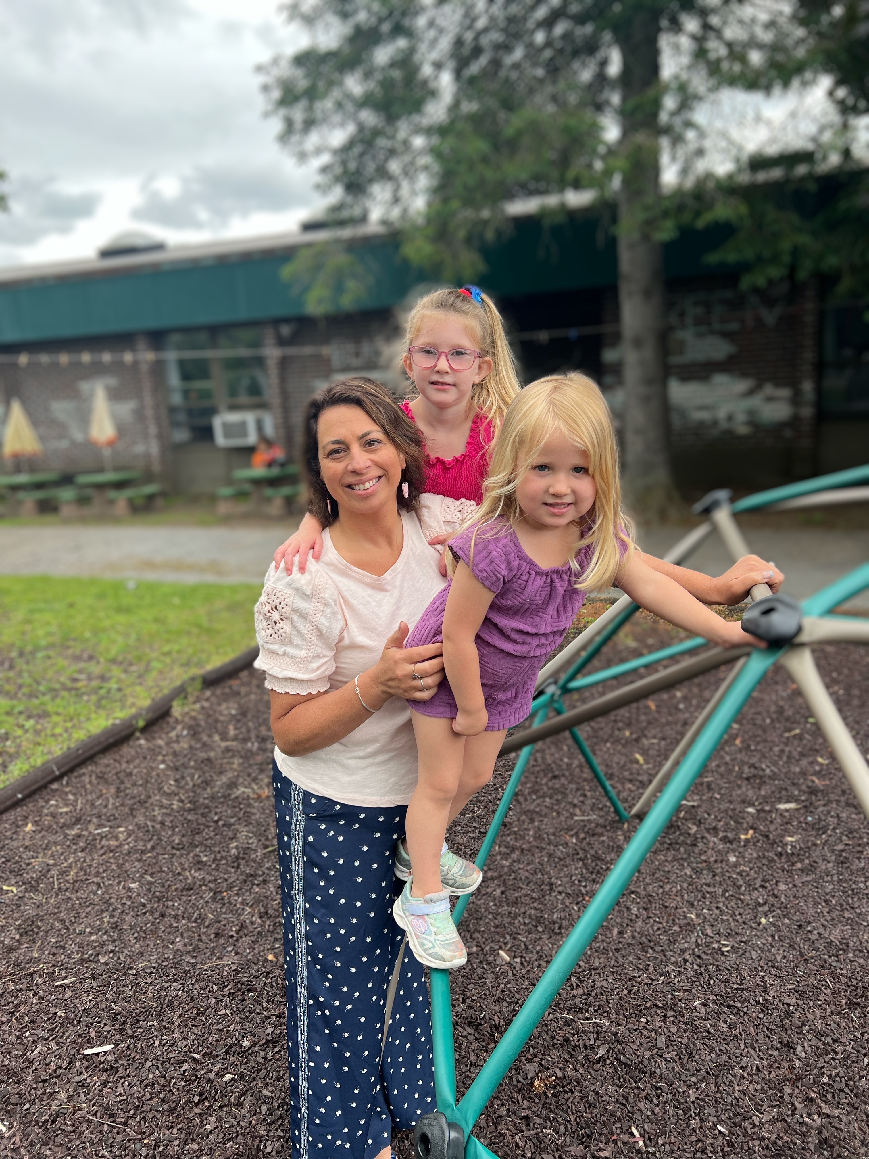 A pale-skinned woman holds two small blonde children who are climbing on a play dome in a playground.