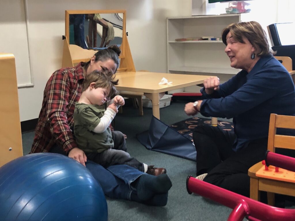 Two pale-skinned women sit on the floor with a toddler on one of their laps. The other woman is caught mid-gesture as she sings to the toddler.