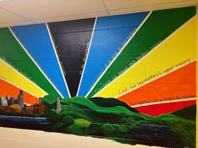 A mural of a rainbow decorated with inspirational sayings, over a picturesque set of rolling hills and a cityscape.