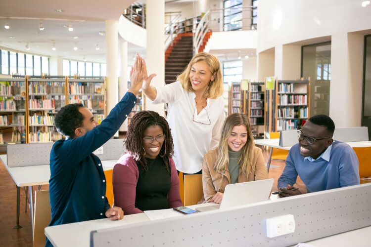 A golden-skinned woman with wavy blonde hair high-fives a Black adult learner seated at a table with other learners