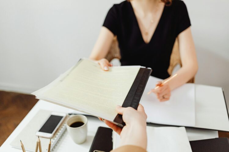 a person off-camera hands a file to a person seated behind a desk. The desktop is already covered by paper files