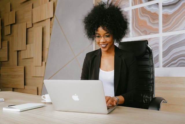 A Black woman with natural hair worn free around her head, sits smiling in front of a laptop