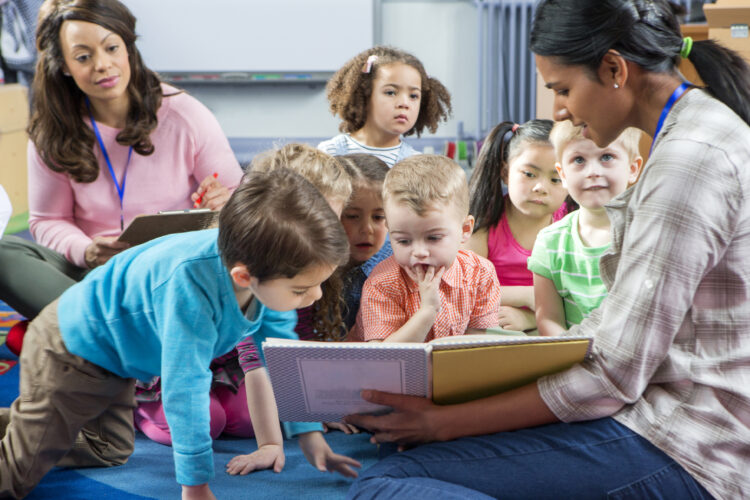 A teacher guides toddlers through story time in a circle, while another teacher evaluates her in the background