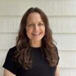 Smiling woman with long wavy brown hair wearing a black short-sleeved top, standing in front of a white textured wall with horizontal siding.