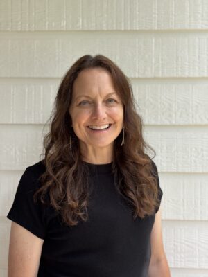 Smiling woman with long wavy brown hair wearing a black short-sleeved top, standing in front of a white textured wall with horizontal siding.