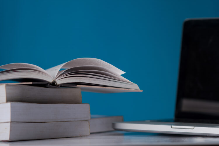 Open book on a stack of closed books next to a laptop on a desk with a blue background.
