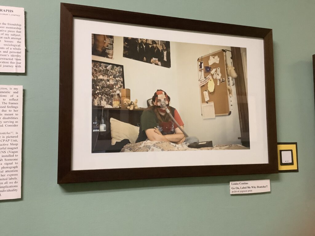 framed photo of a young pale-skinned woman sitting on a bed wearing a CPAP mask. The photo has a credit on the wall: "Louise Contino. 'Go On, Label Me, Why Dontcha?!" Archival print."