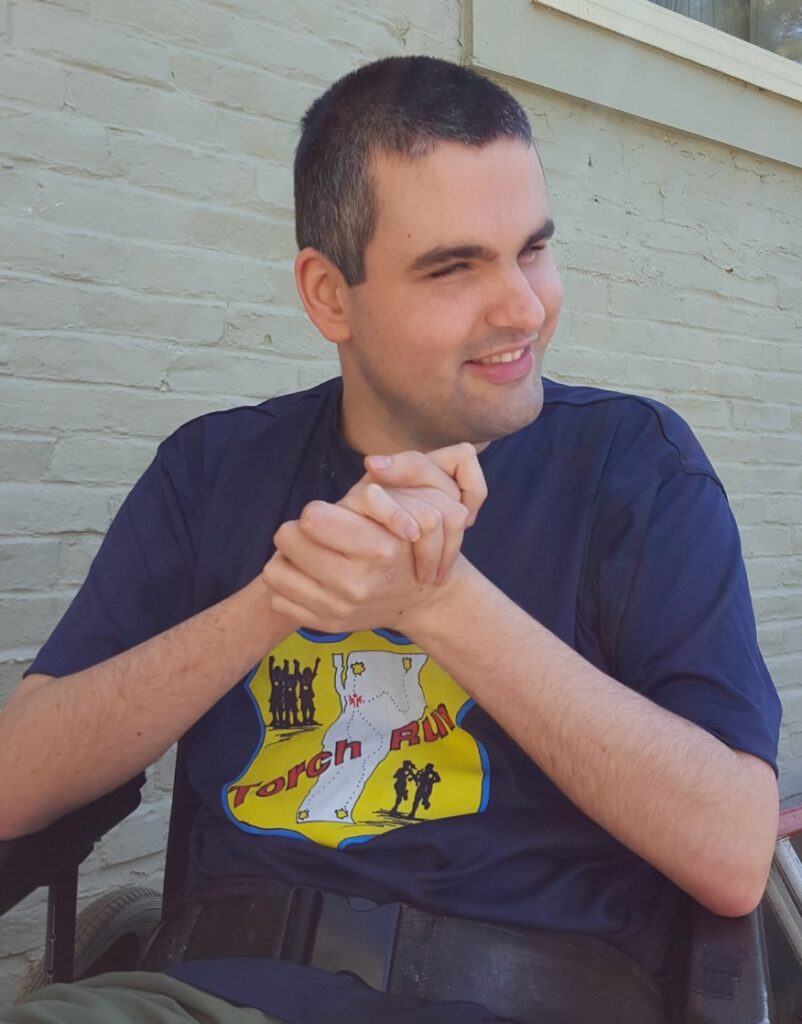 A pale-skinned non-visual man with short dark hair flecked with gray clasps his hands together in front of him