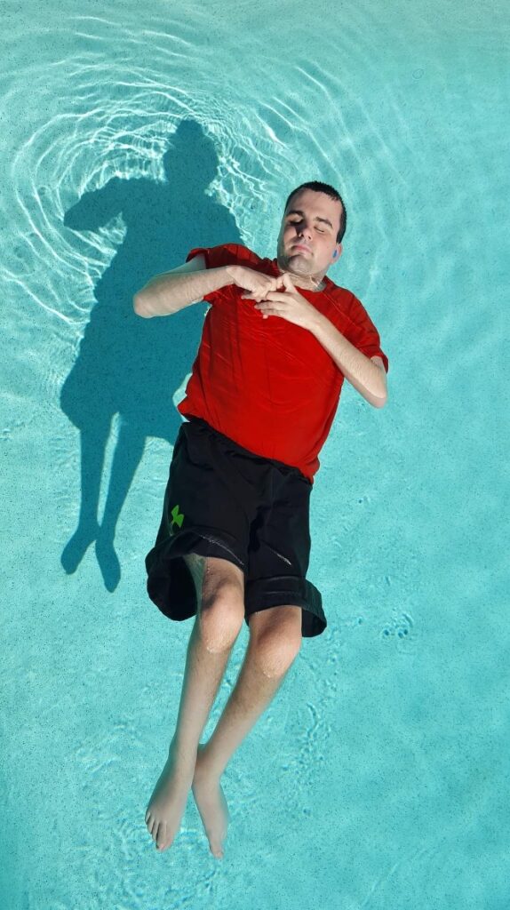 A pale-skinned man in his mid-twenties, with short dark brown hair, floating on his back in a pool, knees tucked up and hands clasped on his chest.