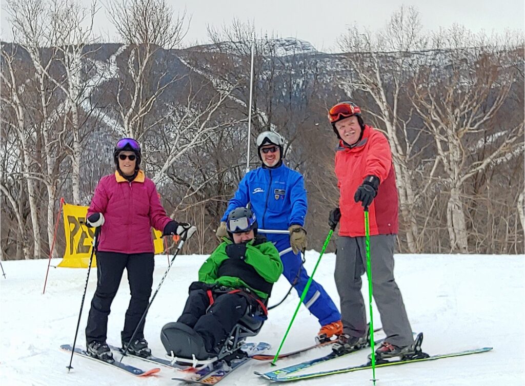 Four adults pose for a photo on skis, in front of stark trees and a snowy mountainside. One of the adults sits in an adaptive ski chair.