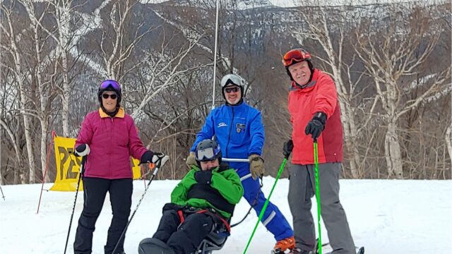 Four adults pose for a photo on skis, in front of stark trees and a snowy mountainside. One of the adults sits in an adaptive ski chair.