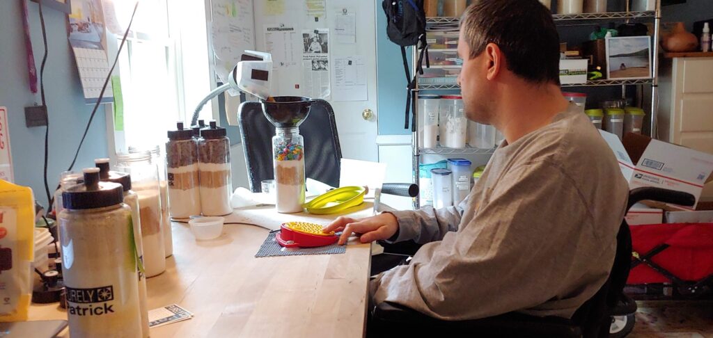 A pale-skinned man in his early thirties sits in a wheelchair at a wooden table, operating a switch. The switch pours layers of candy into a clear sports bottle.