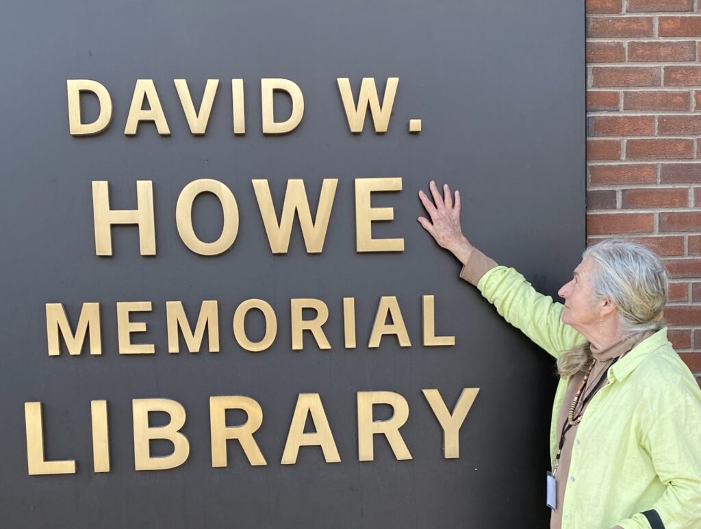 A pale-skinned Indigenous older woman with gray hair pulled back in a bun rests a hand on a large gray wall plaque. Picked out in gold letters on the plaque: David. W. Howe Memorial Library.