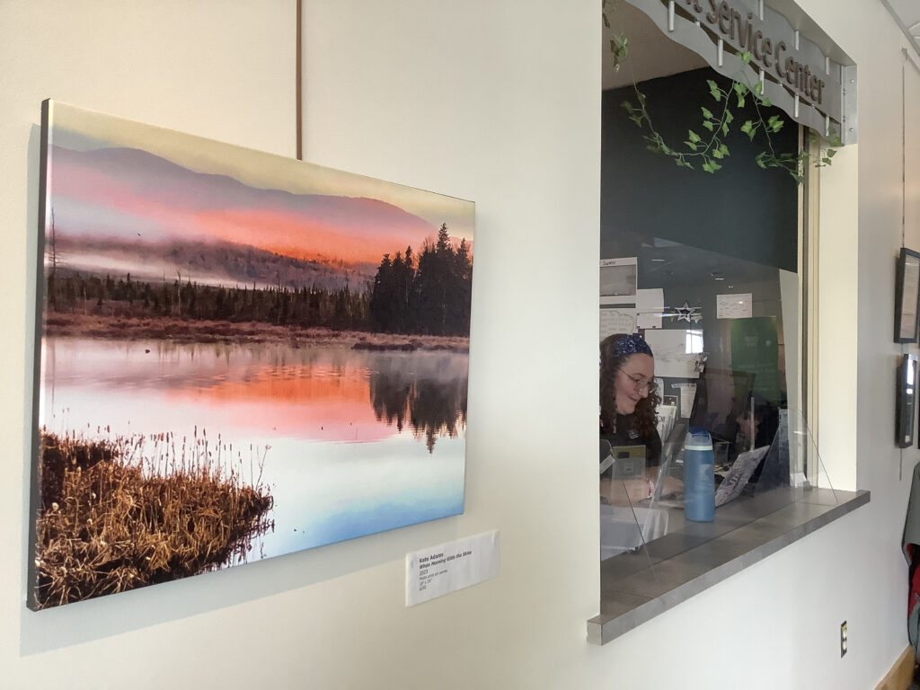 A large photograph of a stunning sunrise over marshes hangs next to an information window, at which sits a college-aged worker.