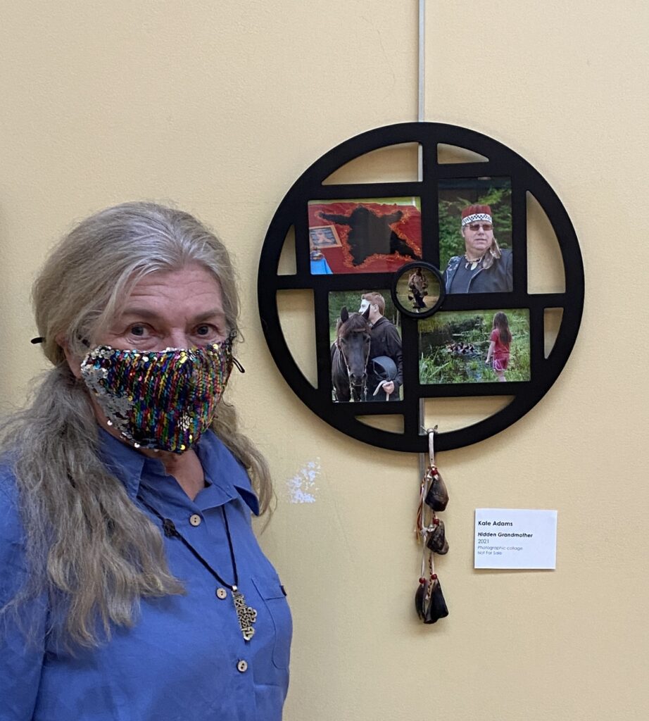 A pale-skinned Indigenous older woman with gray hair pulled back in a low ponytail stands wearing a sequined medical mask next to a circular piece of art on a wall. The art features horses, a child, a man in Indigenous dress, and a bearskin rug.