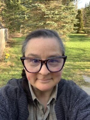 a white non-binary person in their mid-forties with dark hair in a braid over one shoulder, wearing large, heavy-framed glasses, and looking quizzically at the camera