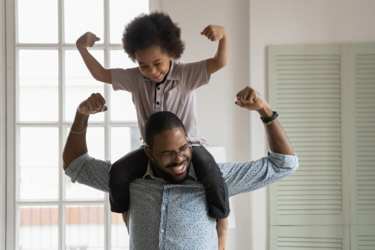 A Black father holds his Black young son on his shoulders, and both flex their muscles in play