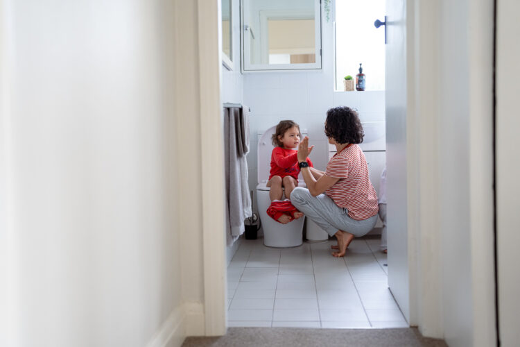 Wide shot of a three year old Eurasian girl sitting on the toilet in the bathroom at home and giving her mom a high five as she provides support and assistance.