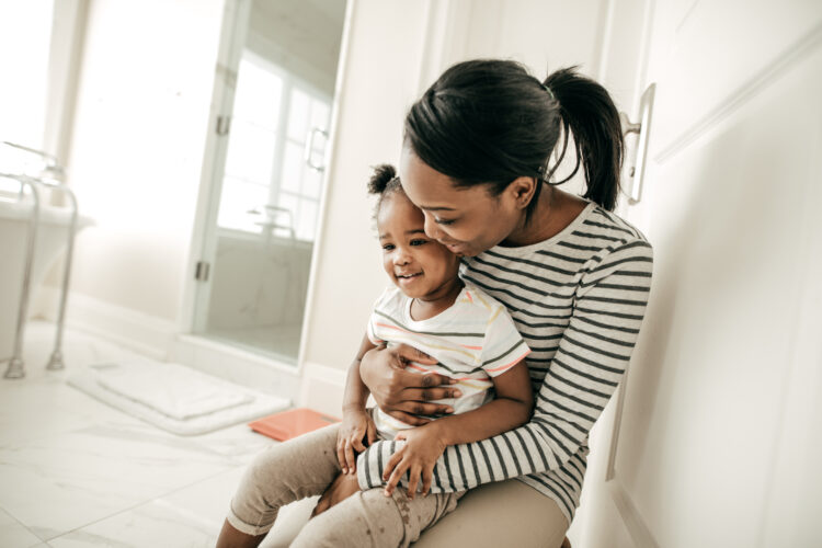 A Black woman holds a Black toddler on her lap, lovingly. The toddler is wearing a tshirt and diapers.