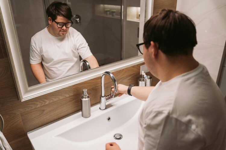 A pale-skinned man in his twenties with Down Syndrome washes his hands at a clean white sink