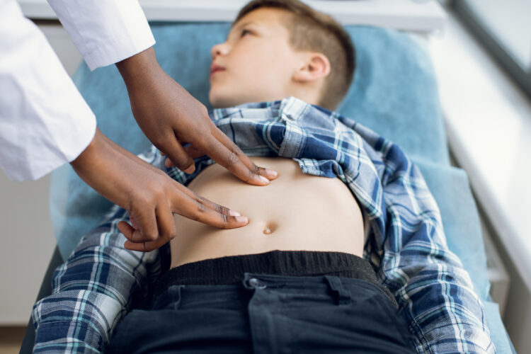 A pale-skinned pre-teen boy lies on an exam bed as a Black doctor presses on his bare stomach.