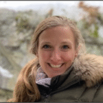 A blonde woman with a ponytail smiling. Wearing a olive colored coat with a furry trim