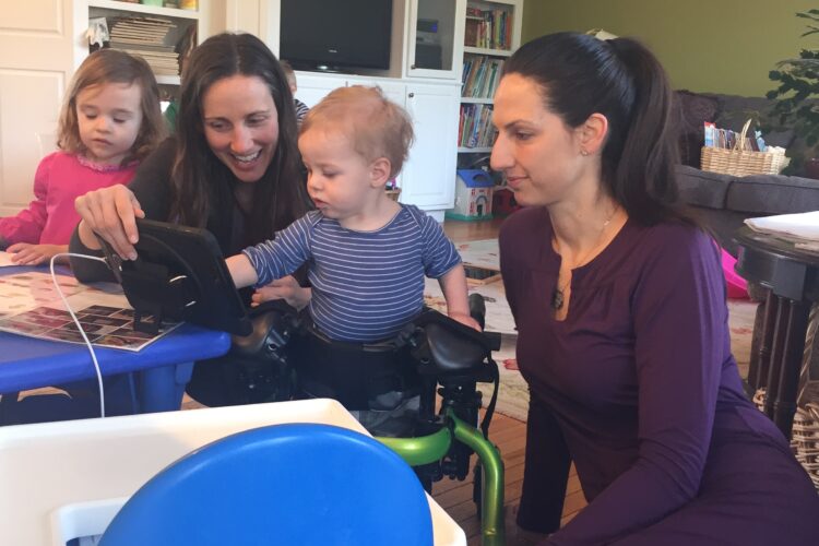 A pale-skinned woman helps a toddler with an AAC device as another toddler and another woman look on.