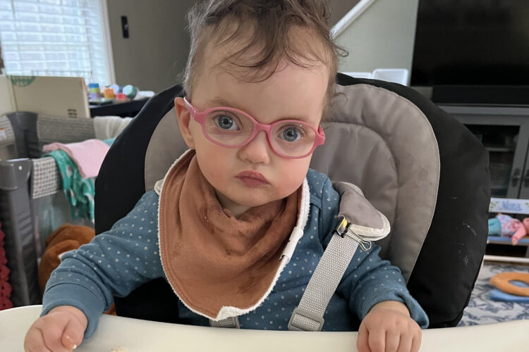 A toddler with pale skin and dark brown curls and tiny pink glasses looks balefully at the camera from a high chair.