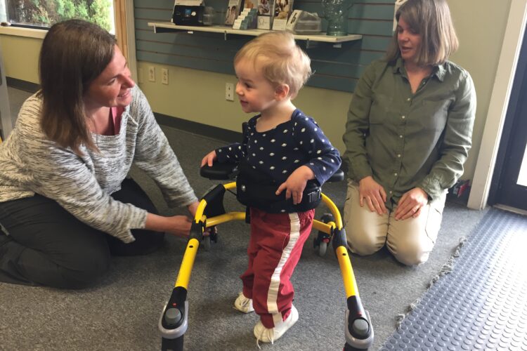 A middle-aged white woman supports a blond toddler in a gait trainer, as another white woman looks on