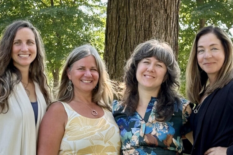 A quartet of women pose next to a tree, smiling.