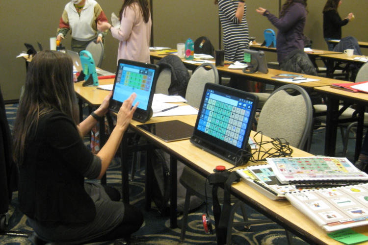 A woman with long dark straight hair kneels in front of an iPad mounted on a table, interacting with the screen while other people converse in the background