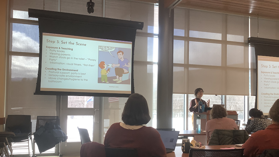 A middle-aged white woman lectures to a packed conference room. To her right is a large projection screen showing a cartoon about toilet training