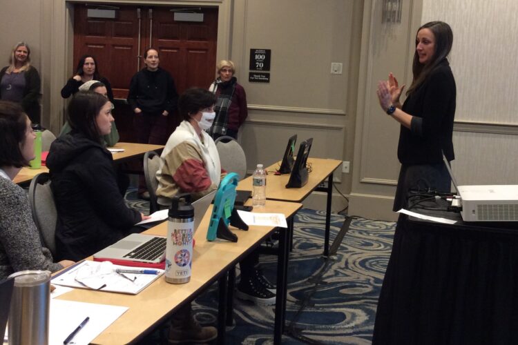 A pale-skinned woman with long dark hair lectures at the front of a conference room