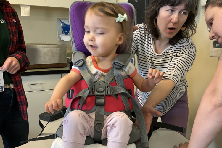 A group of adults adjust a supportive chair containing a small blonde toddler girl