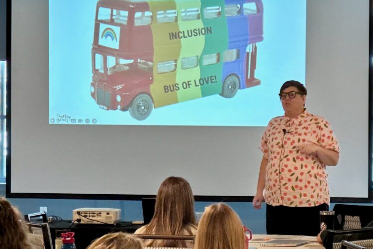 A white woman in her thirties with short brown hair and glasses stands in front of a projector screen. on the screen is a rainbow bus.