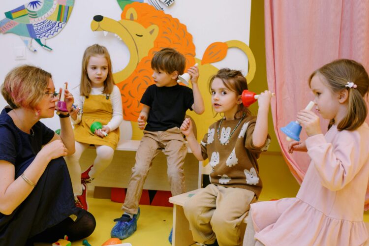 A teacher kneels with several students in a classroom as they hold toys to their ears