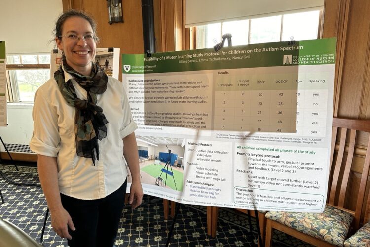 A pale-skinned woman in her forties, with dark hair pulled back into a low ponytail and thin-rimmed glasses, poses for a photo next to a research poster