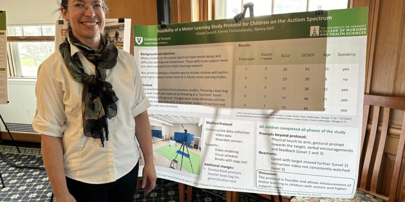 A pale-skinned woman in her forties, with dark hair pulled back into a low ponytail and thin-rimmed glasses, poses for a photo next to a research poster