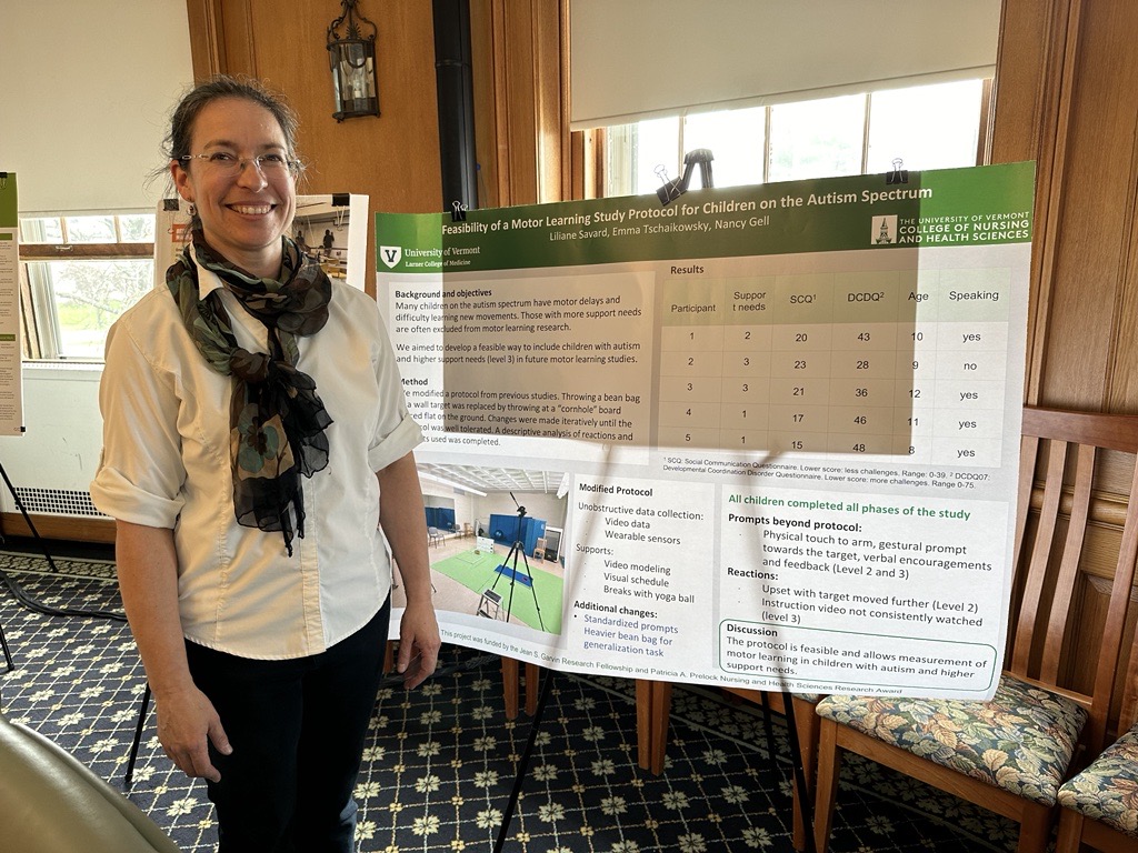 A pale-skinned woman in her forties, with dark hair pulled back into a low ponytail and thin-rimmed glasses, poses for a photo next to a research poster