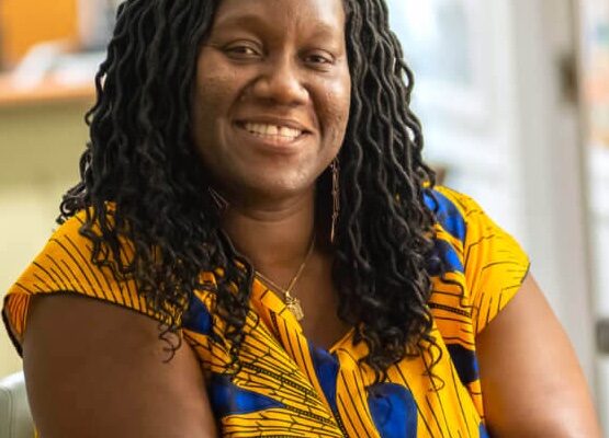 A Black woman in her forties with hair in loose, curled locs to her shoulders sits smiling, in a gold and cobalt short-sleeved top