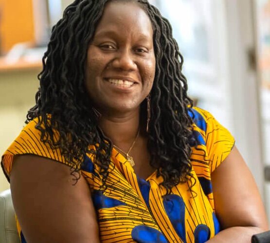 A Black woman in her forties with hair in loose, curled locs to her shoulders sits smiling, in a gold and cobalt short-sleeved top