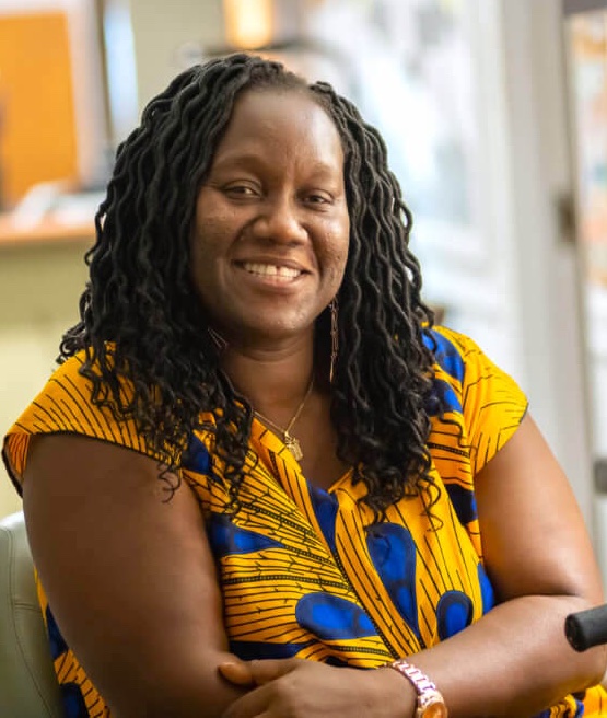 A Black woman in her forties with hair in loose, curled locs to her shoulders sits smiling, in a gold and cobalt short-sleeved top
