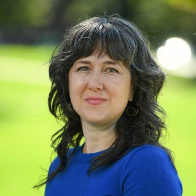 Chayah Lichtig: a white woman in her late thirties with wavy dark hair worn with bangs, in a royal blue shirt, poses for a photo outside