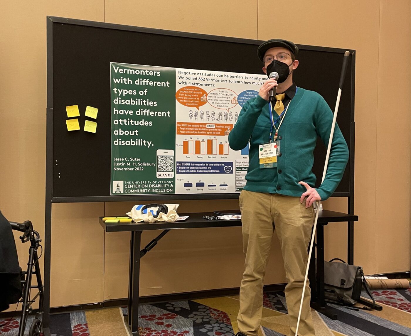 A masked blind man in a flat cap with a white cane speaks into a microphone in front of a research poster